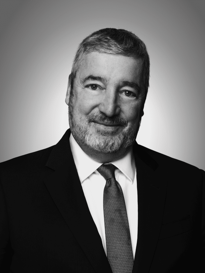 A middle-aged man with short gray hair and a beard, wearing a suit and tie, poses against a plain gradient background.