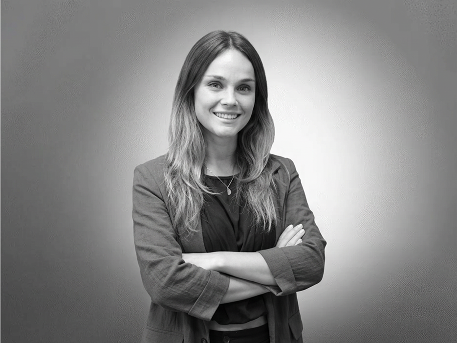 Woman with long hair in a blazer stands with arms crossed, smiling at the camera against a plain grey background.