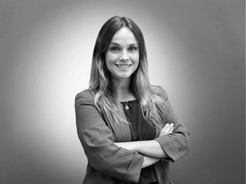 Woman with long hair stands smiling, arms crossed, wearing a blazer. The background is a plain, gradient grey.